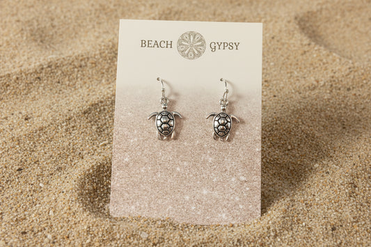 Turtle-shaped earrings on a 'Beach Gypsy' branded card against a sand ackground
