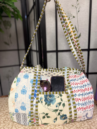 A handmade patchwork fabric shoulder bag with green and white striped borders, floral blue patterns, and red embroidered details. The bag is shown holding a smartphone and sunglasses, set against a background of a black screen and a beige textured surface.