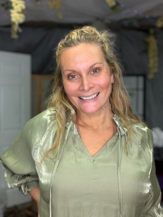 A smiling blonde woman wearing a shiny sage green satin blouse, photographed indoors against a dark draped backdrop with hanging floral decorations.