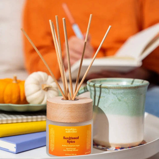 A reed diffuser bottle labeled 'Sun-Kissed Spice' with natural wooden reeds, placed on a table beside a ceramic mug, books, and small pumpkins, with a person in an orange sweater blurred in the background reading a book.