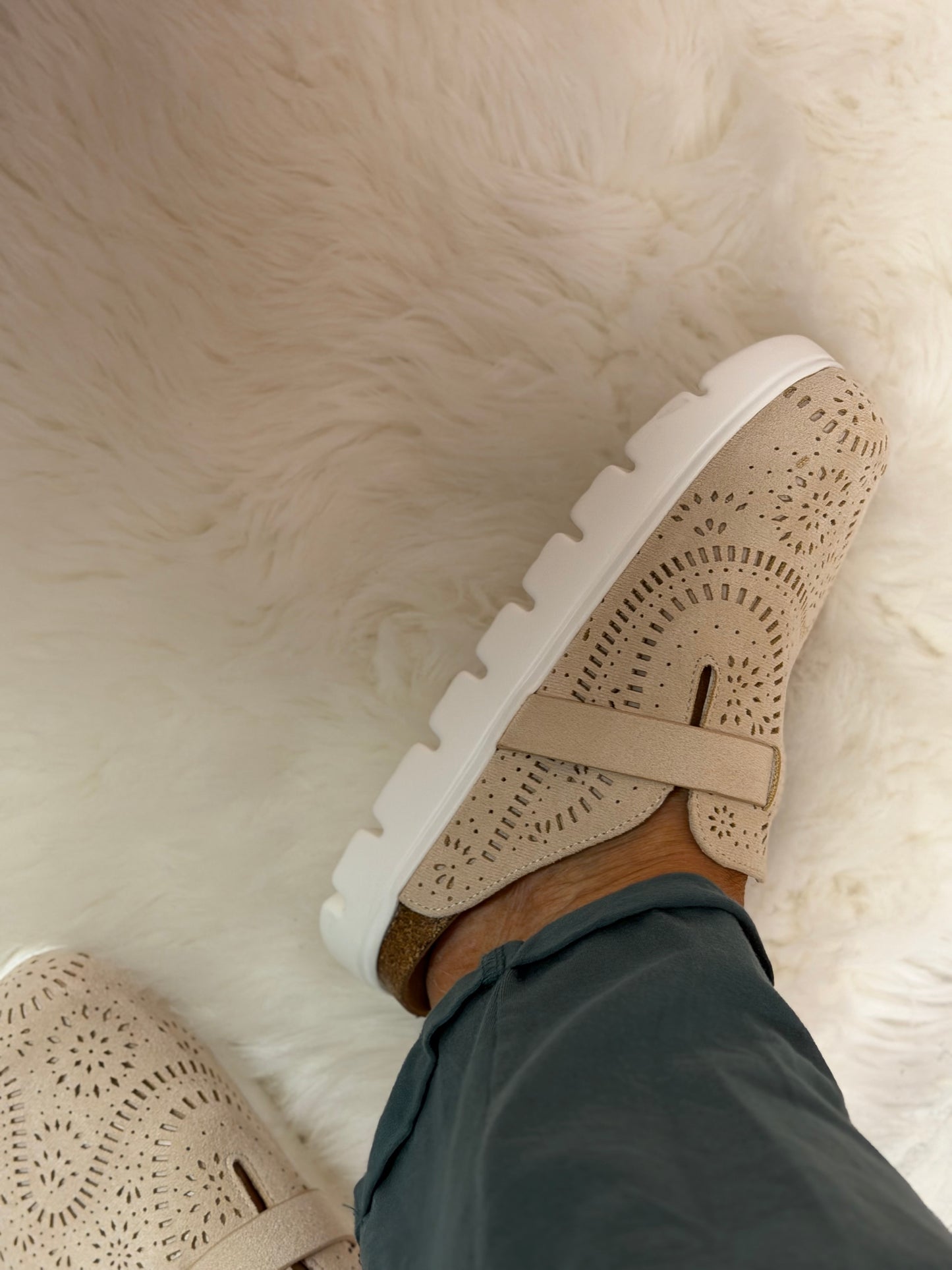 A close-up of a person wearing a beige perforated clog-style shoe with a white sole, standing on a white faux fur rug. The person is also wearing dark teal pants.
