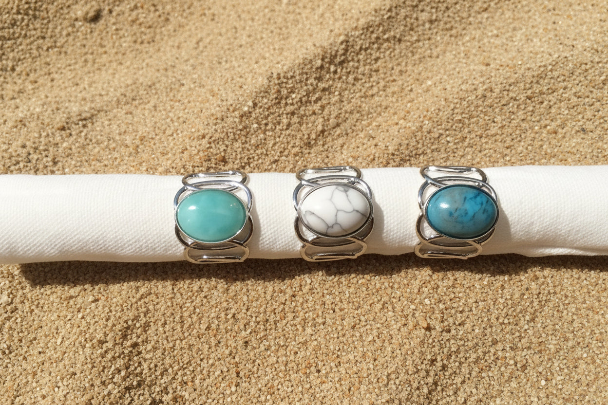 Three silver rings with oval turquoise and marbled stone cabochons displayed on a white fabric on a sand surface 