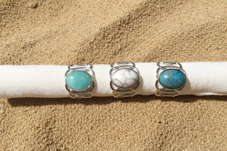Three silver rings with oval turquoise and marbled stone cabochons displayed on a white fabric on a sand surface 