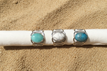Three silver rings with oval turquoise and marbled stone cabochons displayed on a white fabric on a sand surface 