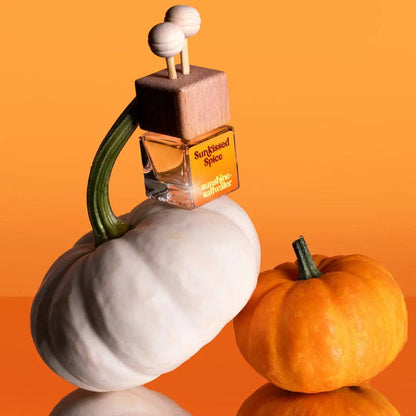 A perfume bottle labeled 'Sunfaded Spice' with a wooden stopper and two round knobs, styled on top of a white pumpkin next to a small orange pumpkin against a solid orange background.