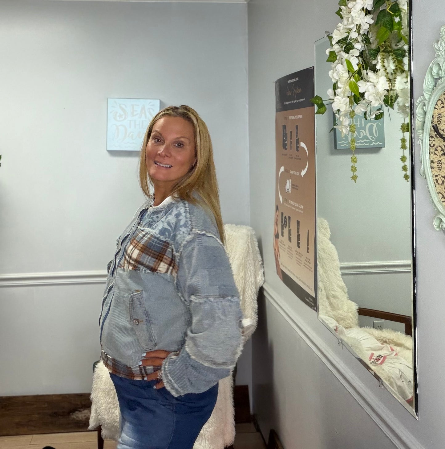 A woman posing in a light blue denim jacket with a patchwork design featuring plaid and faded denim panels, paired with blue jeans, standing in an indoor setting with a mirror and wall decor.