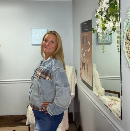 A woman posing in a light blue denim jacket with a patchwork design featuring plaid and faded denim panels, paired with blue jeans, standing in an indoor setting with a mirror and wall decor.