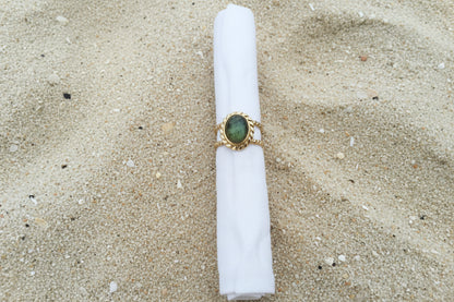 A gold ring with an oval green stone, displayed on a white fabric ring holder against a textured sand background.