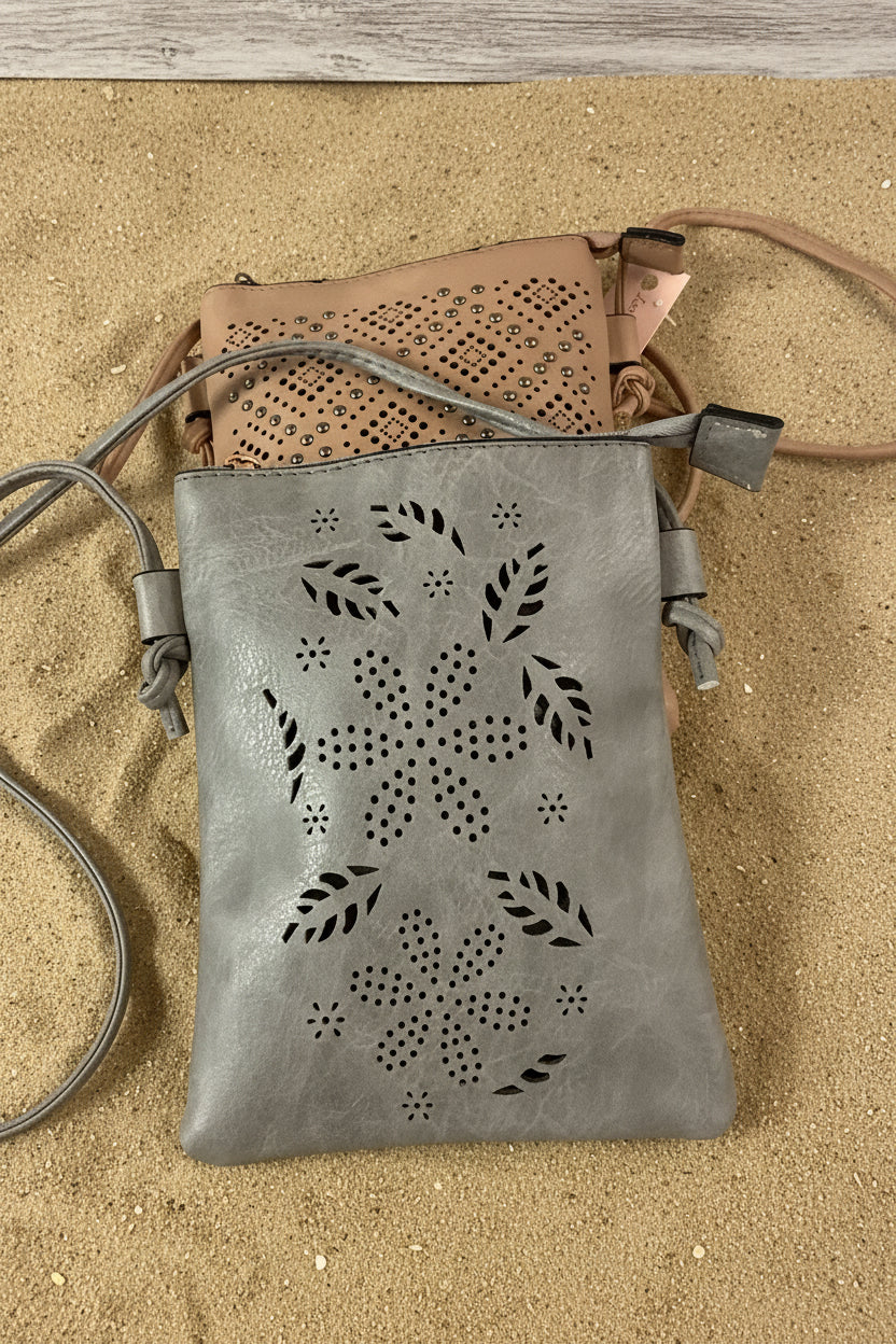 A gray faux-leather crossbody bag with laser-cut floral and leaf patterns, shown with a tan bag partially visible behind it on a bright pink background.