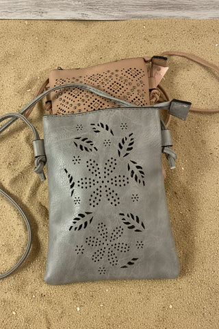 A gray faux-leather crossbody bag with laser-cut floral and leaf patterns, shown with a tan bag partially visible behind it on a bright pink background.
