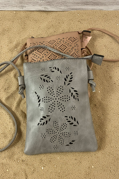 A gray faux-leather crossbody bag with laser-cut floral and leaf patterns, shown with a tan bag partially visible behind it on a bright pink background.