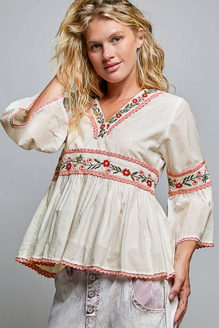 A woman wearing a cream-colored bohemian-style blouse with red and green floral embroidery, paired with light-wash denim jeans, against a solid gray studio background.