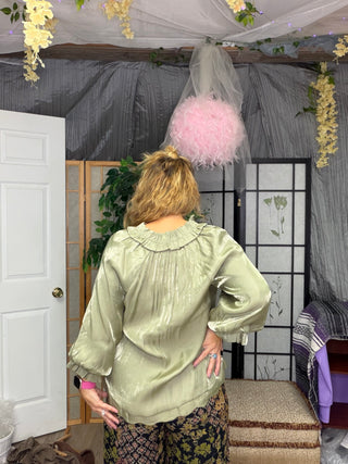 A woman viewed from behind wearing a shiny sage green blouse with a ruffled collar and voluminous sleeves, standing in a decorated room with a gray curtain backdrop, floral garlands, and a pink feather lamp.