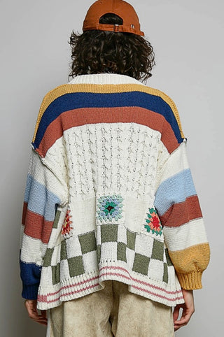 A person shown from the back wearing a colorful striped and patterned knit cardigan, a brown cap, and light beige pants against a plain light gray studio background.