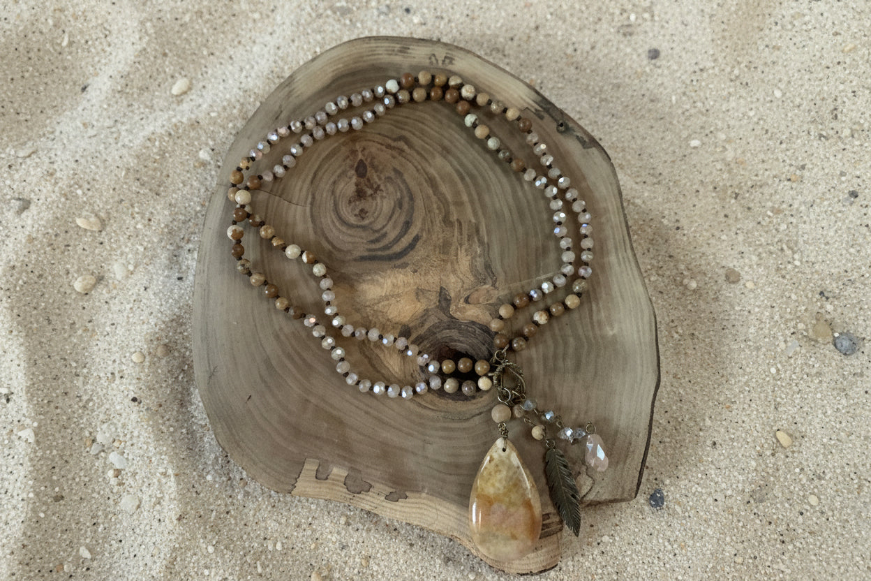 A beaded necklace with earthy brown, beige, and gray beads, featuring a large polished tan stone pendant and a feather accent, displayed on a rustic wood slice over a sandy background.