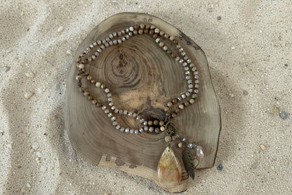 A beaded necklace with earthy brown, beige, and gray beads, featuring a large polished tan stone pendant and a feather accent, displayed on a rustic wood slice over a sandy background.