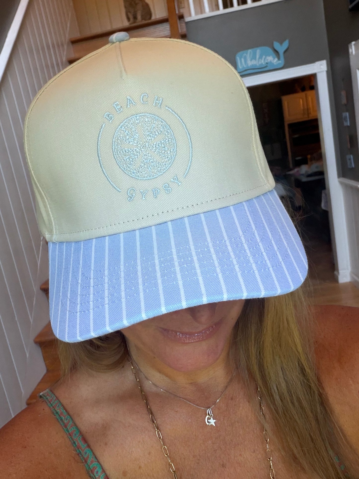 A woman wearing a Beach Gypsy brand baseball cap with a pale yellow crown and a light blue and white striped visor, photographed indoors.