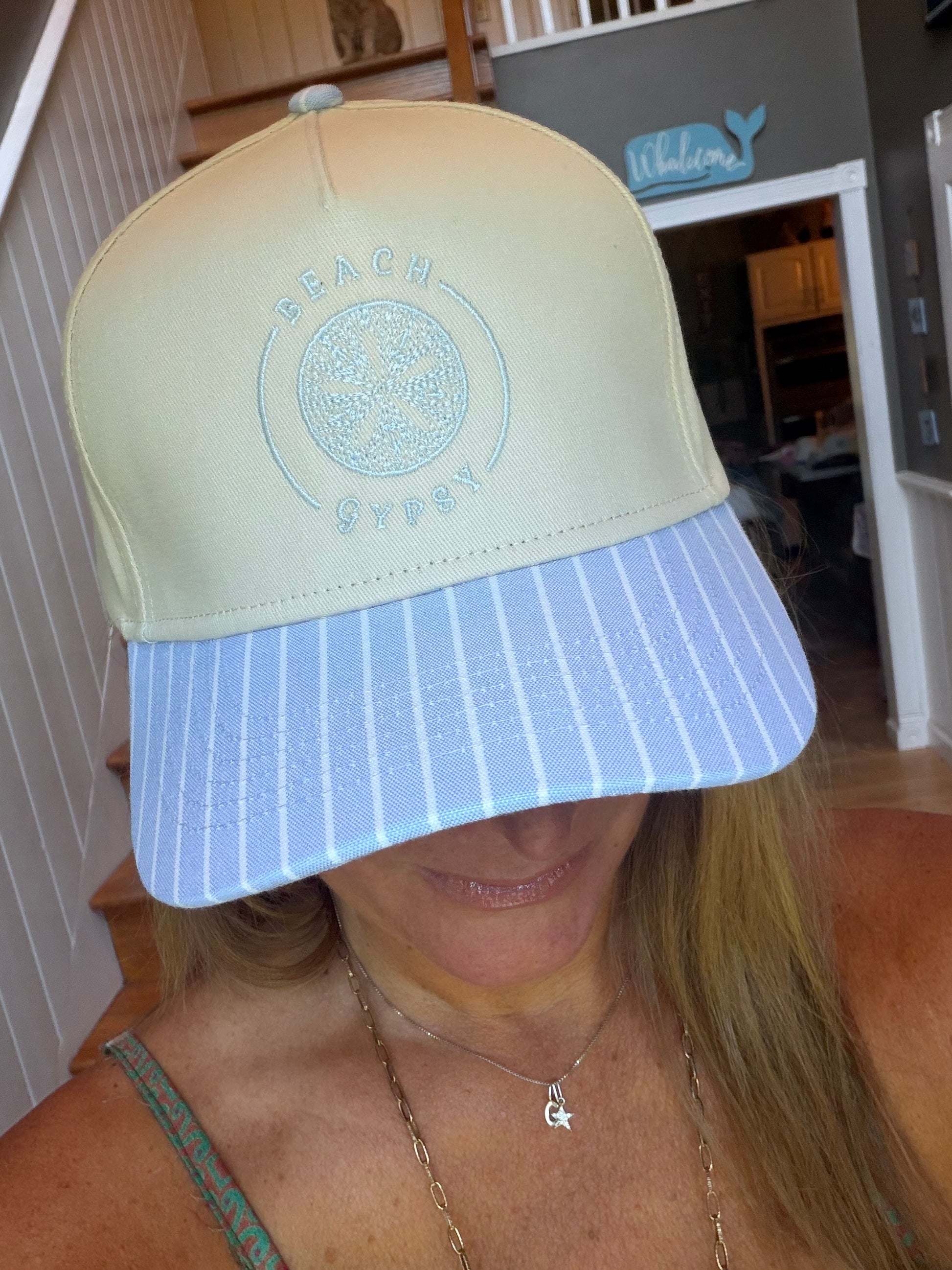 A woman wearing a Beach Gypsy brand baseball cap with a pale yellow crown and a light blue and white striped visor, photographed indoors.