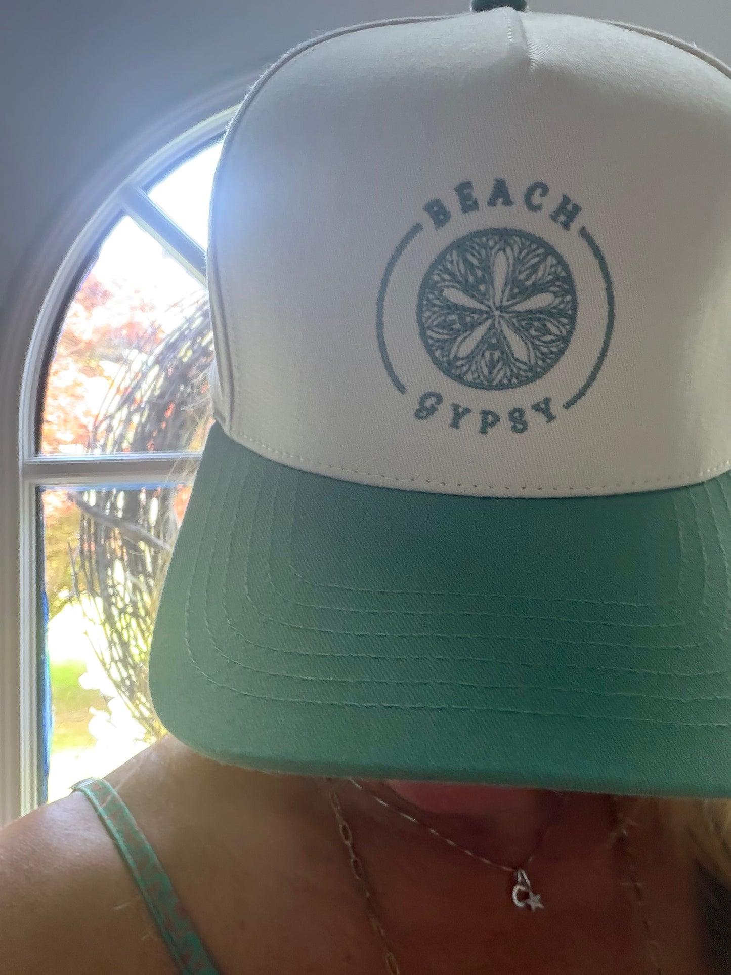 A close-up of a beautiful woman wearing a two-tone trucker hat with a white crown and a mint green brim. The front of the hat features a dark green circular logo with a starfish design and the words 'BEACH GYPSY'.
