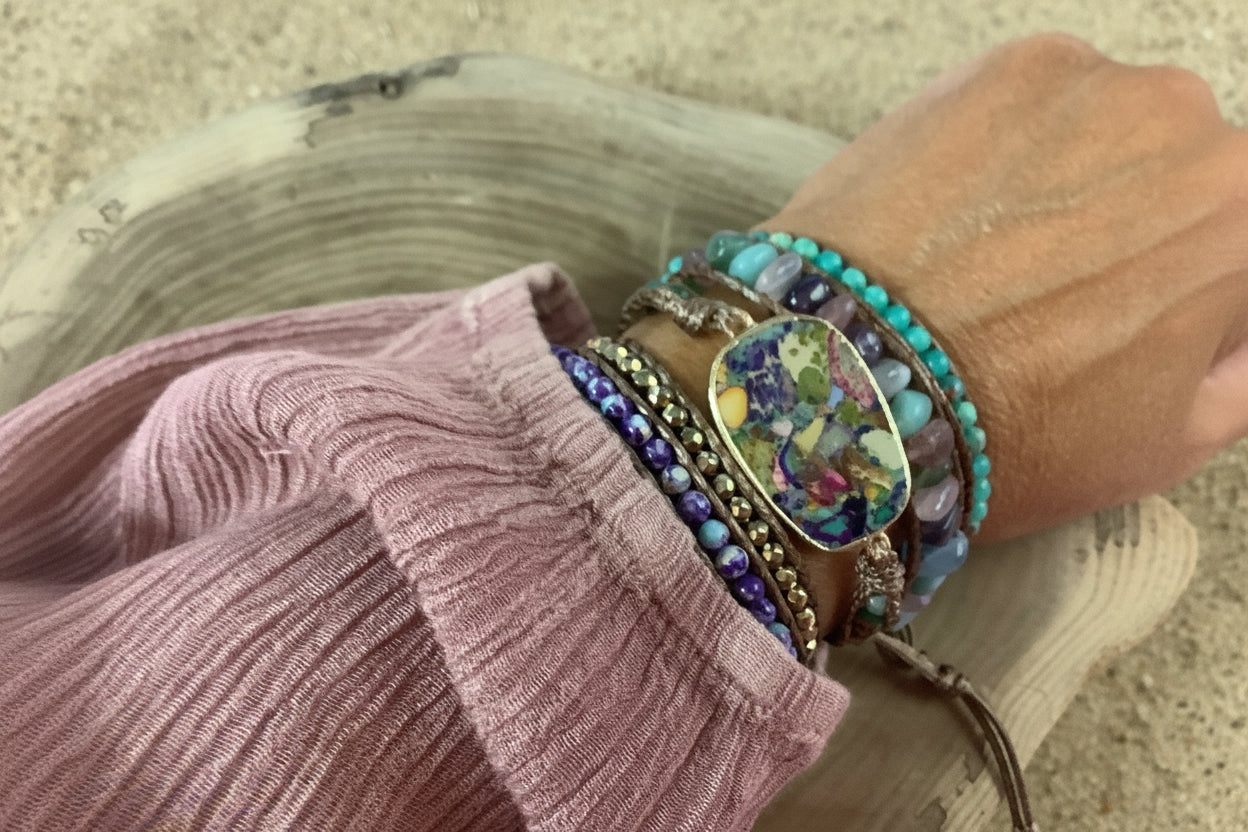 A close-up of a person's wrist wearing multiple colorful beaded bracelets and a central square charm bracelet with a multicolor floral inlay, styled over a dusty rose sleeve on a wooden surface against a sand background.