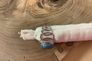 A handmade silver ring with a blue marbled stone and a wrapped silver coil, displayed on a white fabric sleeve over a rustic wood slice background.