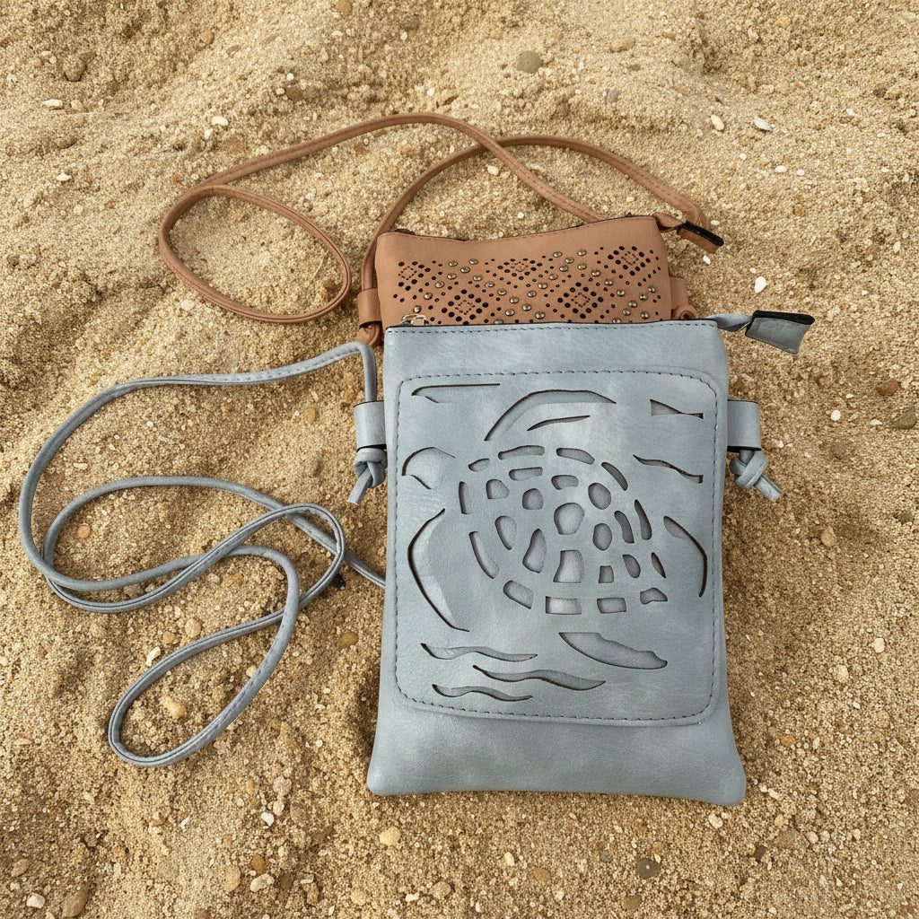 A gray-blue faux-leather crossbody bag with a laser-cut turtle design on the front pocket, shown with its brown perforated top flap and gray strap on a bright pink background.