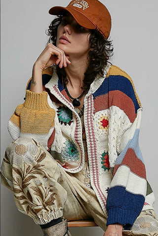 A model wearing a chunky, multicolored patchwork knit cardigan, beige embroidered pants, and an orange baseball cap, posed against a neutral gray background.