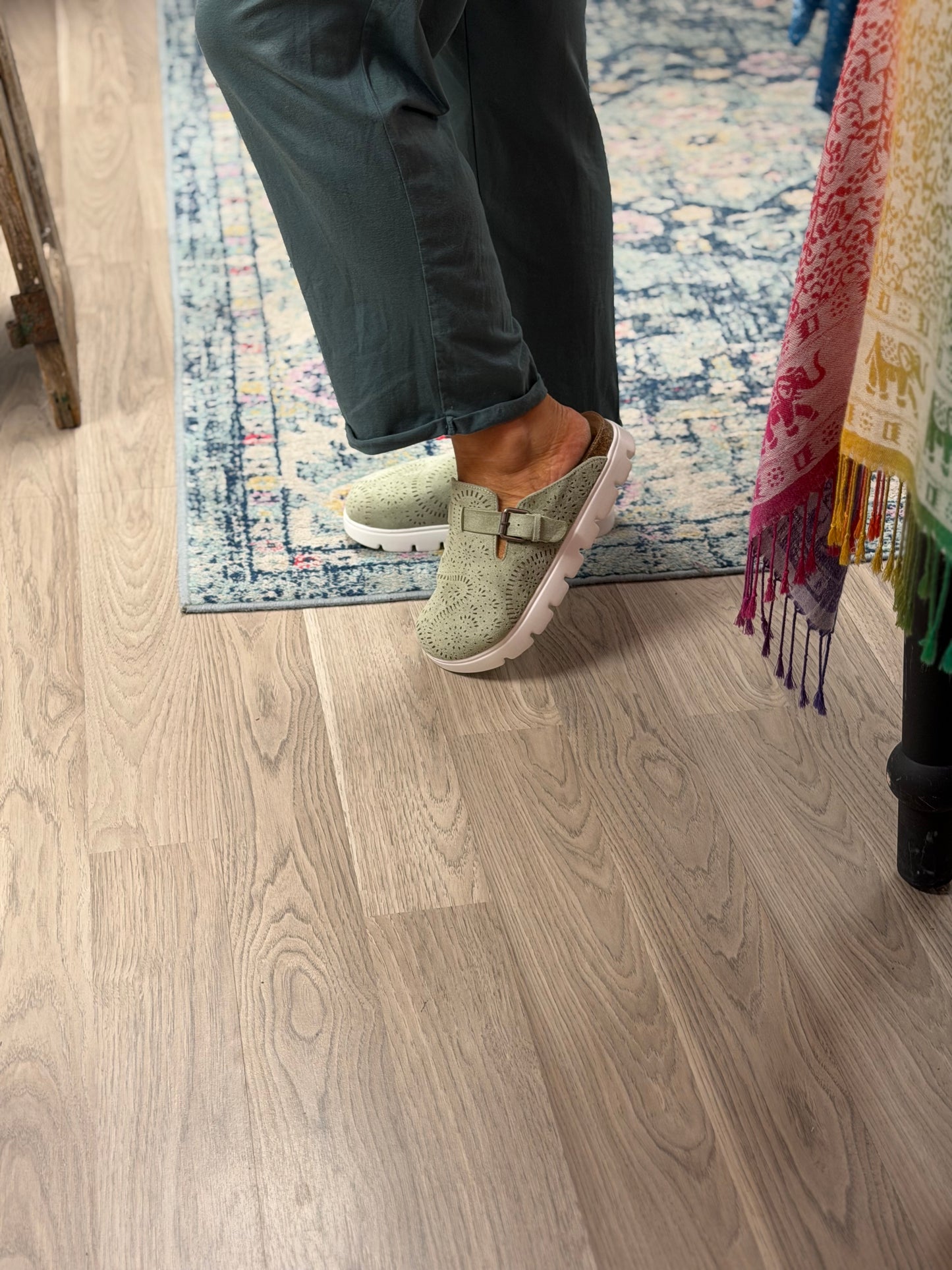 A person wearing light sage green slip-on clogs with a buckle strap, standing on a light gray wood-look floor next to a blue patterned rug.