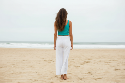 Model walking on beach wearing turquoise tank top and white eyelet pants