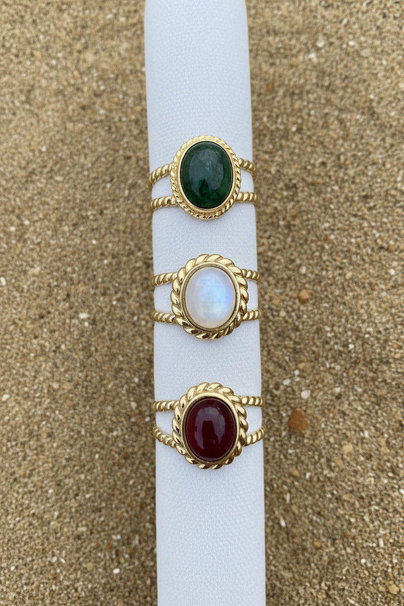 Three gold-toned ring bands displayed vertically on a white cylindrical stand, set against a textured tan woven fabric background. The rings feature oval cabochon stones in green, pink stone and orangish red.