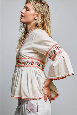 A woman wearing a cream-colored bohemian-style blouse with red and green floral embroidery on the waistband and sleeve cuffs, paired with light gray distressed jeans, photographed against a solid gray studio background.