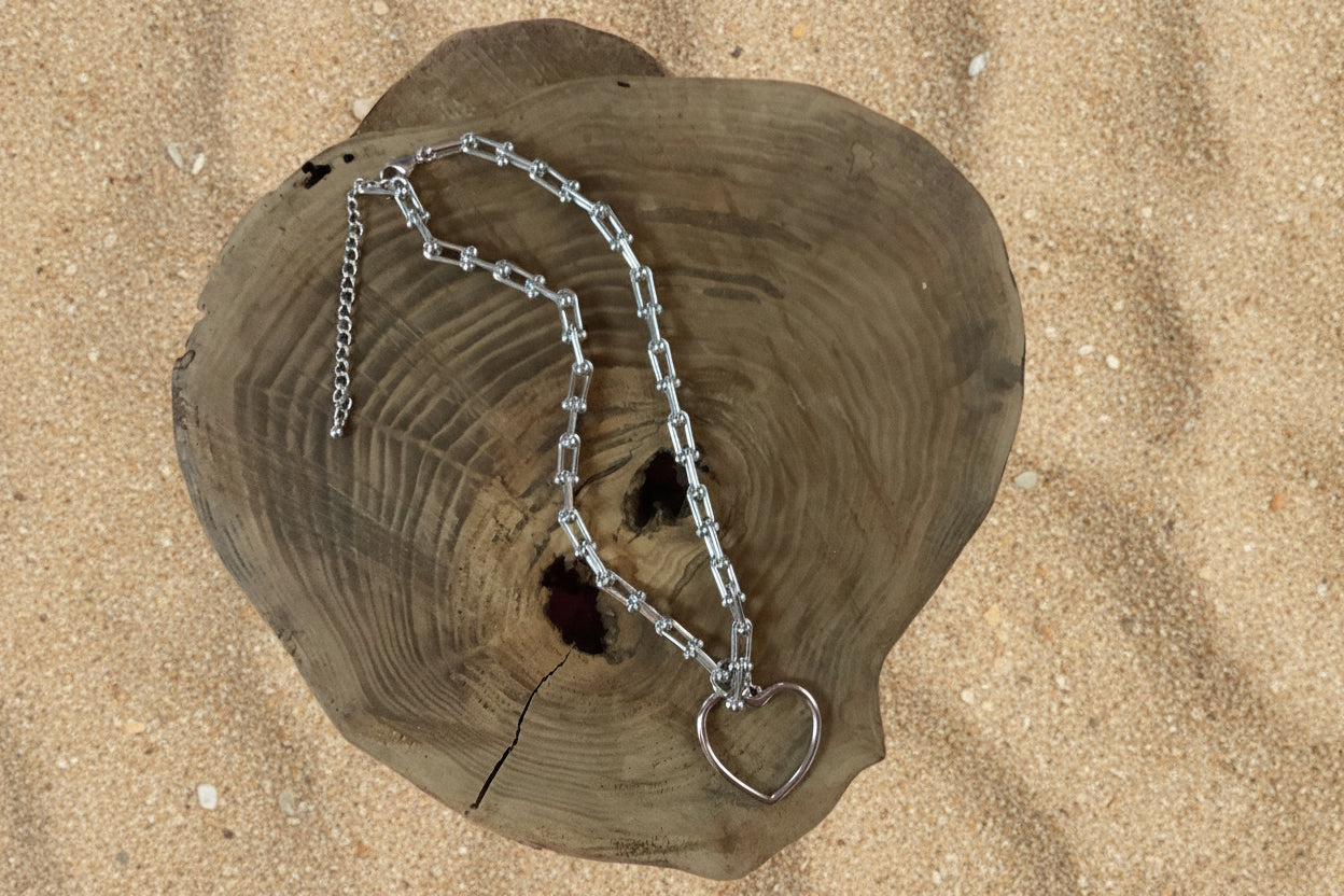 A silver-toned chain necklace with a heart-shaped pendant, displayed on a rustic heart-shaped wooden slice against a sand  background.