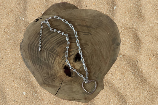 A silver-toned chain necklace with a heart-shaped pendant, displayed on a rustic heart-shaped wooden slice against a sand  background.