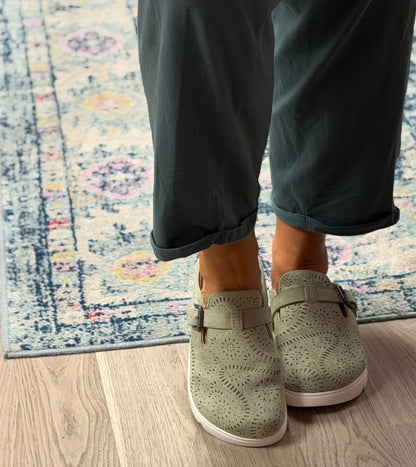 A person wearing sage green perforated slip-on clogs with double straps, standing on a light wood floor next to a patterned area rug.