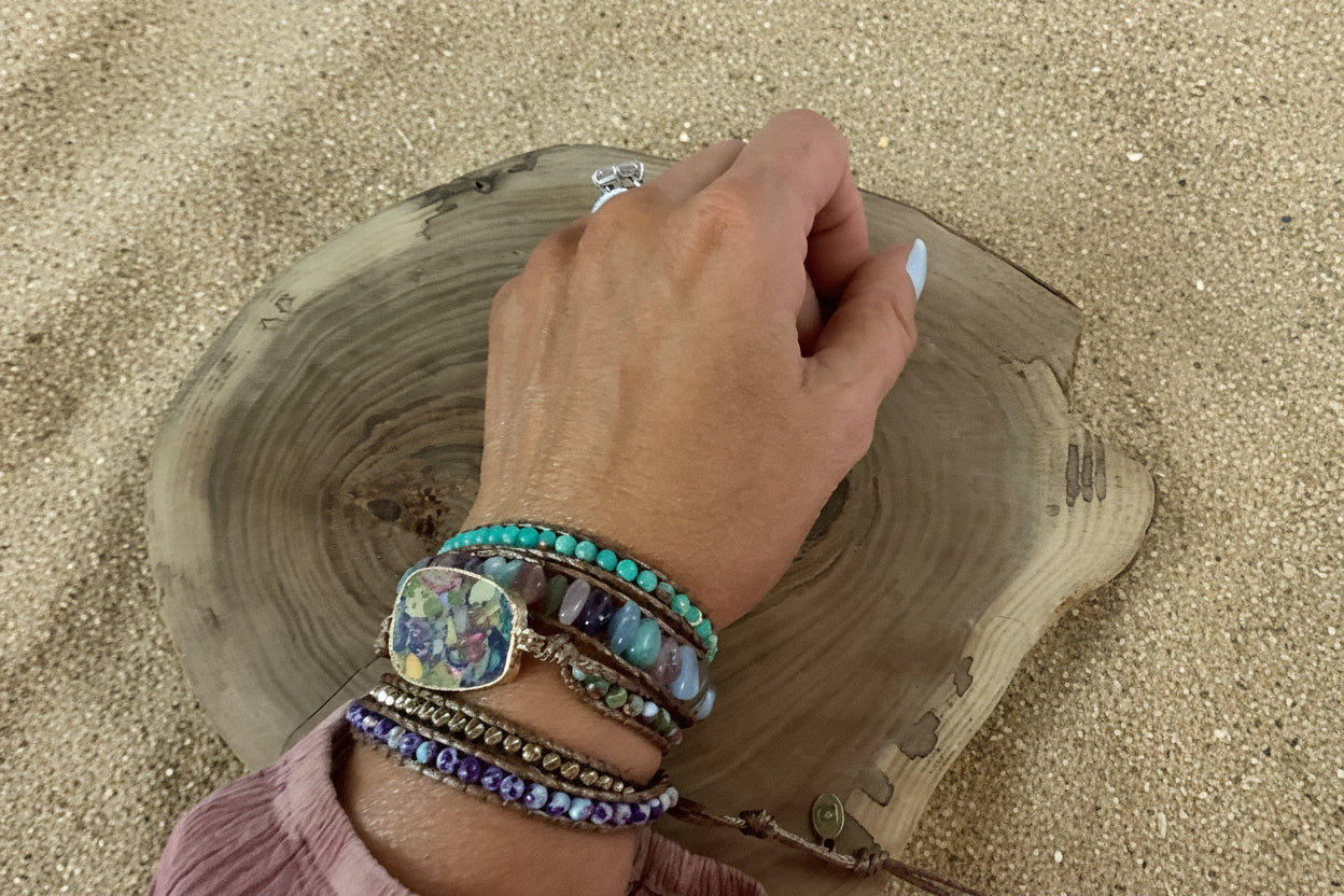 A close-up of a person's hand wearing multiple colorful bracelets, including turquoise, purple, blue, and brown leather bracelets with beads and a floral pendant, resting on a rustic wood slice against a sand background.