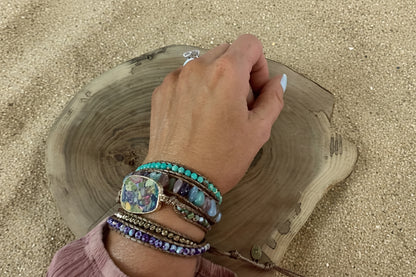 A close-up of a person's hand wearing multiple colorful bracelets, including turquoise, purple, blue, and brown leather bracelets with beads and a floral pendant, resting on a rustic wood slice against a sand background.