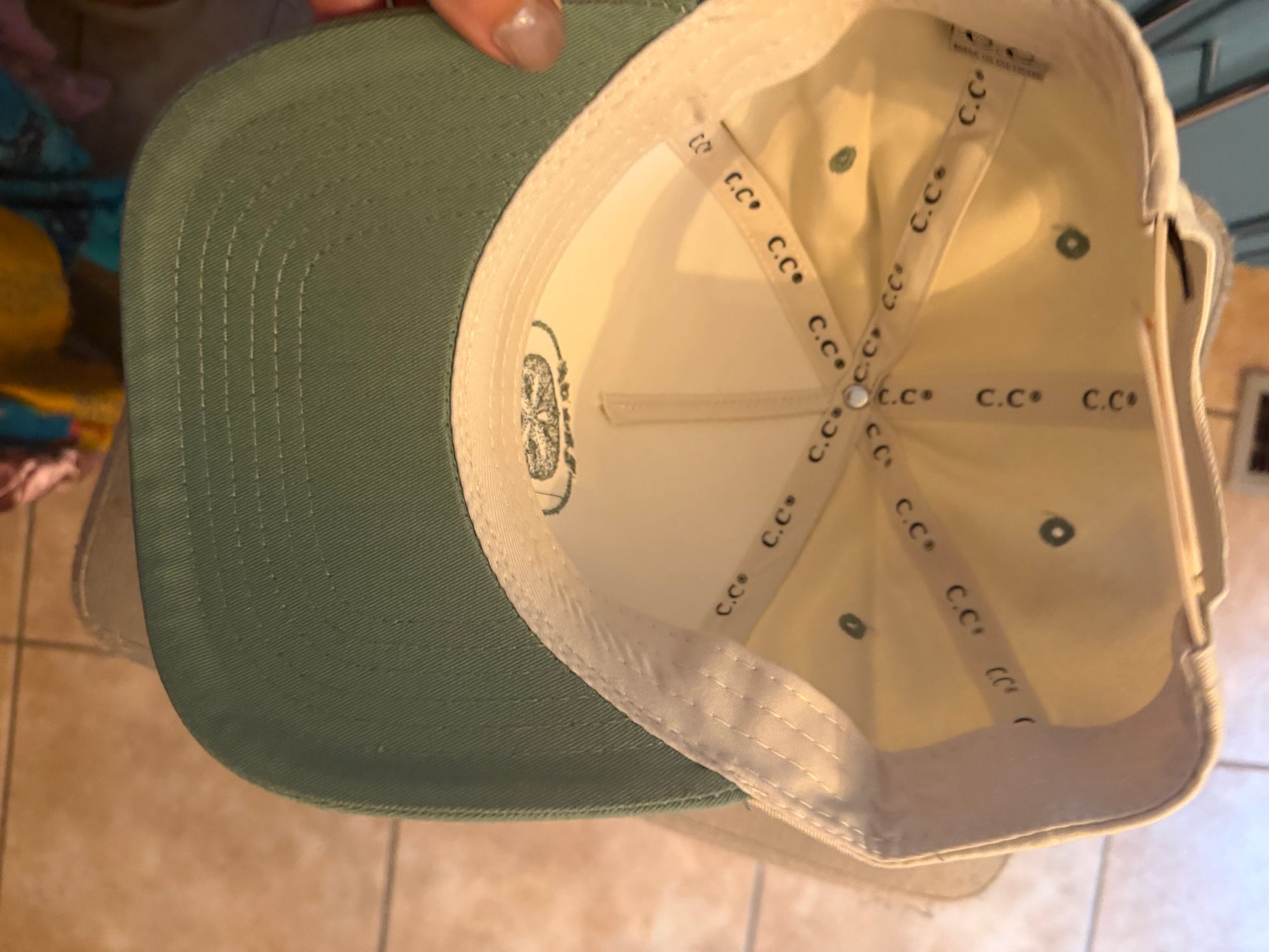A close-up of a baseball cap shown from the inside, featuring an olive green curved brim and a cream-colored interior with 'CC' branding on the panels and a small crest logo on the bill.