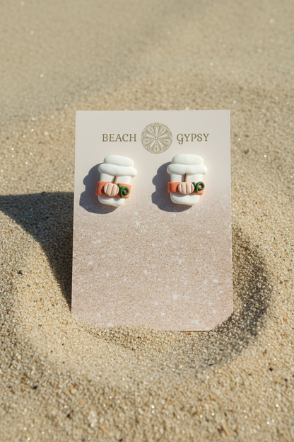 A pair of small stud earrings displayed on a branded card against a sandy beach background. The earrings are white with orange and green details, and the card reads 'BEACH GYPSY'.