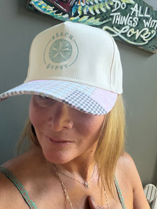 A woman wearing a Beach Gypsy baseball cap in a lifestyle setting, with a gray wall and a decorative wooden sign in the background.