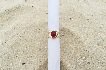 A gold ring with an oval red cabochon gemstone, displayed on a white paper tube against a sand background.