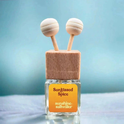 A small glass bottle with a square wooden cap and two round wooden diffuser sticks inserted into the top. The front label reads 'Sunkissed Spice' and 'sunshine-saltwater'.