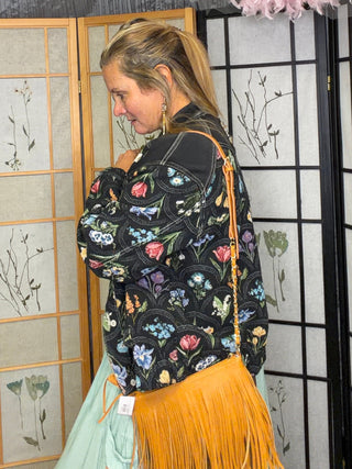 A woman wearing a black floral bomber jacket and a bright orange fringe skirt, standing in front of decorative folding screens.