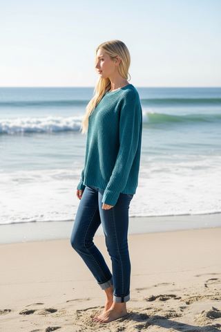 A woman stands on a sandy beach wearing a teal ribbed knit sweater and dark blue jeans, with the ocean waves and pale sky in the background.