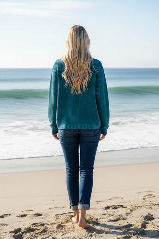 A woman with long blonde hair stands on a sandy beach facing the ocean, wearing a teal knit sweater and dark blue jeans.