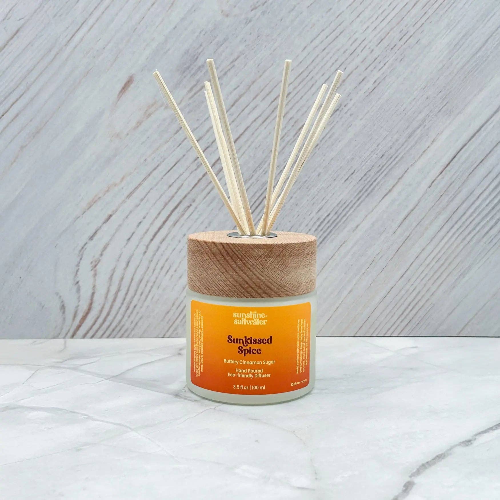 A reed diffuser bottle with a light wood cap and natural reeds, labeled 'Sunblazed Spice' by 'The Smell Store', set on a white marble surface against a light gray wood-grain background.