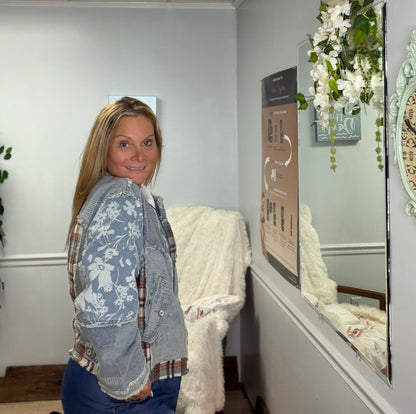 Side view of a woman with blond hair standing in a grey interior room with wall decor. She is wearing a blue denim jacket with patchwork detailing and a zippered front 