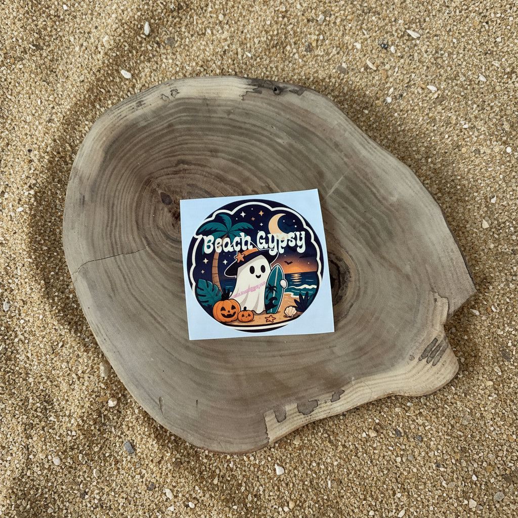 A square sticker with a Halloween beach illustration, placed on a round natural wood slice against a sand background. The sticker features the text 'Beach Gypsy' and depicts a ghost, pumpkins, palm trees, and a crescent moon.