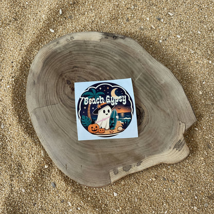 A square sticker with a Halloween beach illustration, placed on a round natural wood slice against a sand background. The sticker features the text 'Beach Gypsy' and depicts a ghost, pumpkins, palm trees, and a crescent moon.