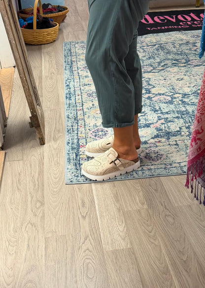 A person wearing beige knit clogs with white soles, standing on a blue and white patterned rug over a light wood floor.