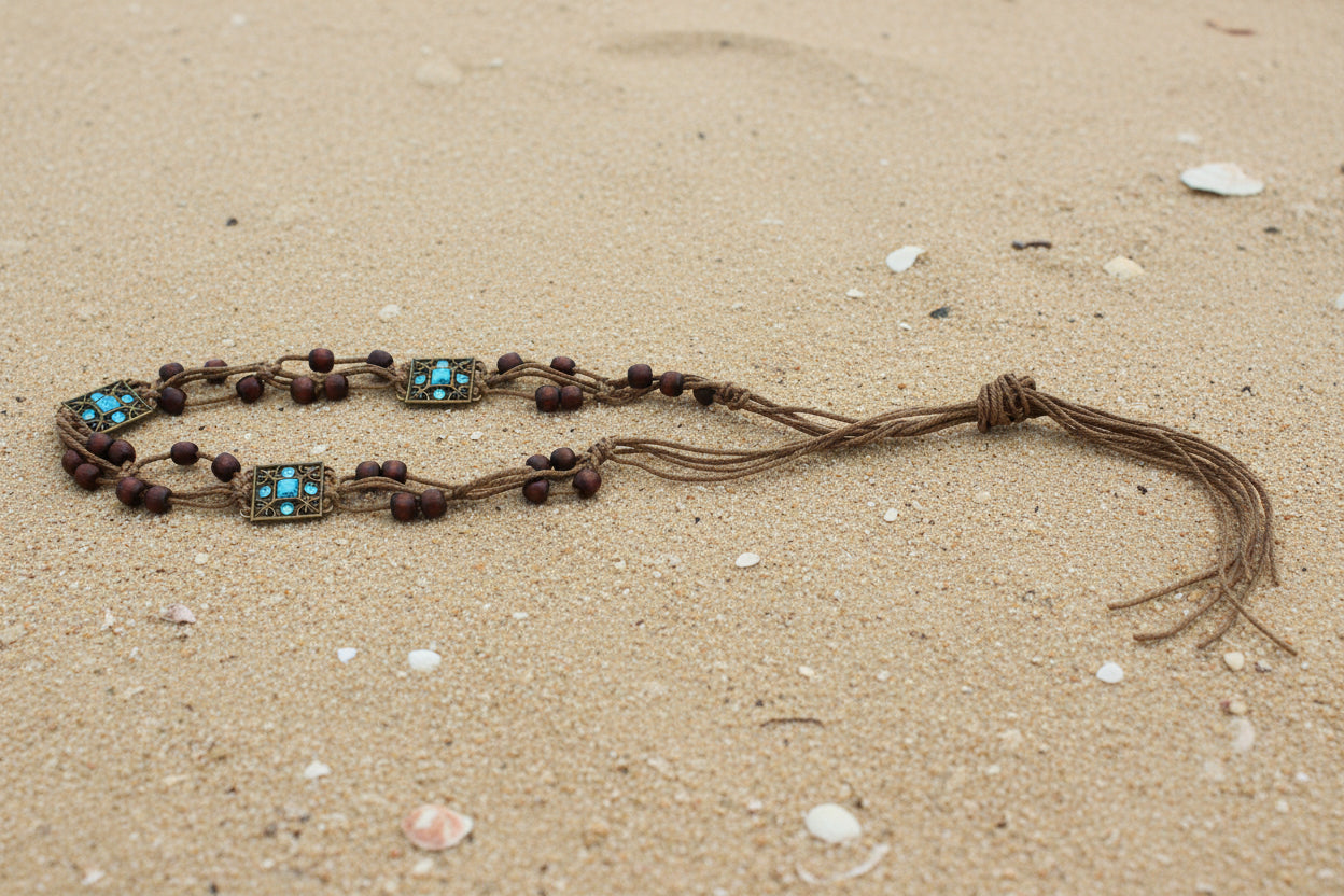 A long brown cord necklace with turquoise and silver-toned rectangular beads and dark brown beads, laid out on a sandy beach 